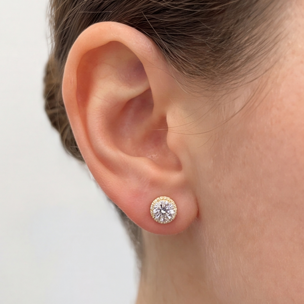 A close-up of a person's ear wearing a small round stud earring with a halo of clear stones and a central clear gemstone in a yellow metal setting.