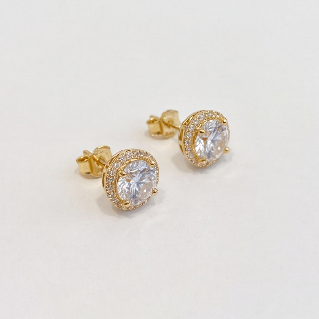 A pair of gold stud earrings with round clear gemstones in halo settings, photographed on a plain light background.