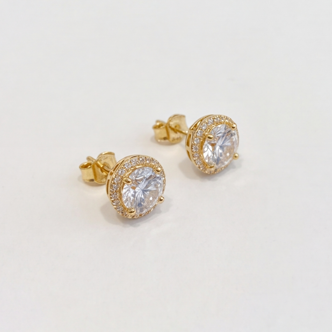 A pair of gold stud earrings with round clear gemstones in halo settings, photographed on a plain light background.