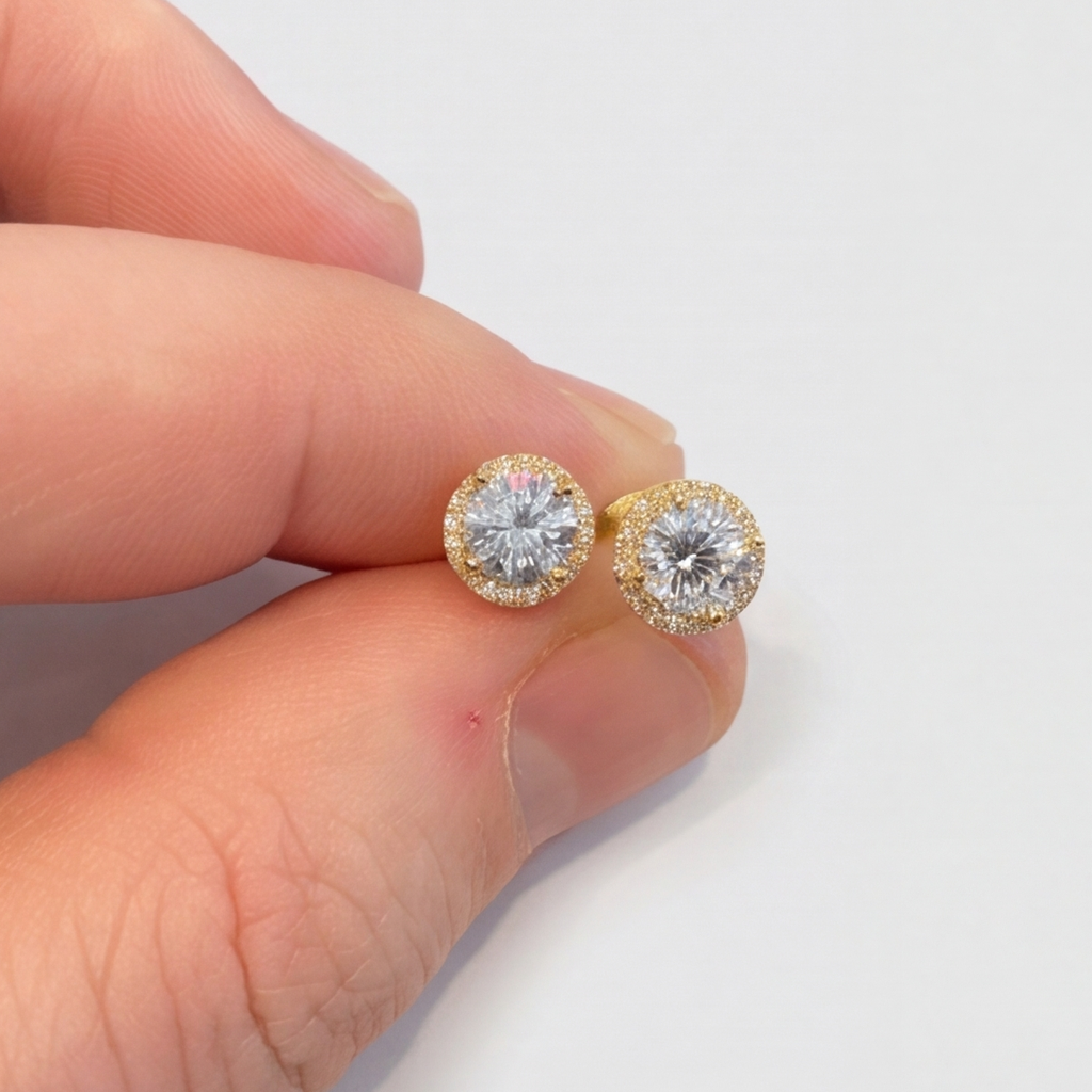A close-up of a person's hand holding a pair of round yellow gold stud earrings with halo settings and clear center stones against a plain white background.