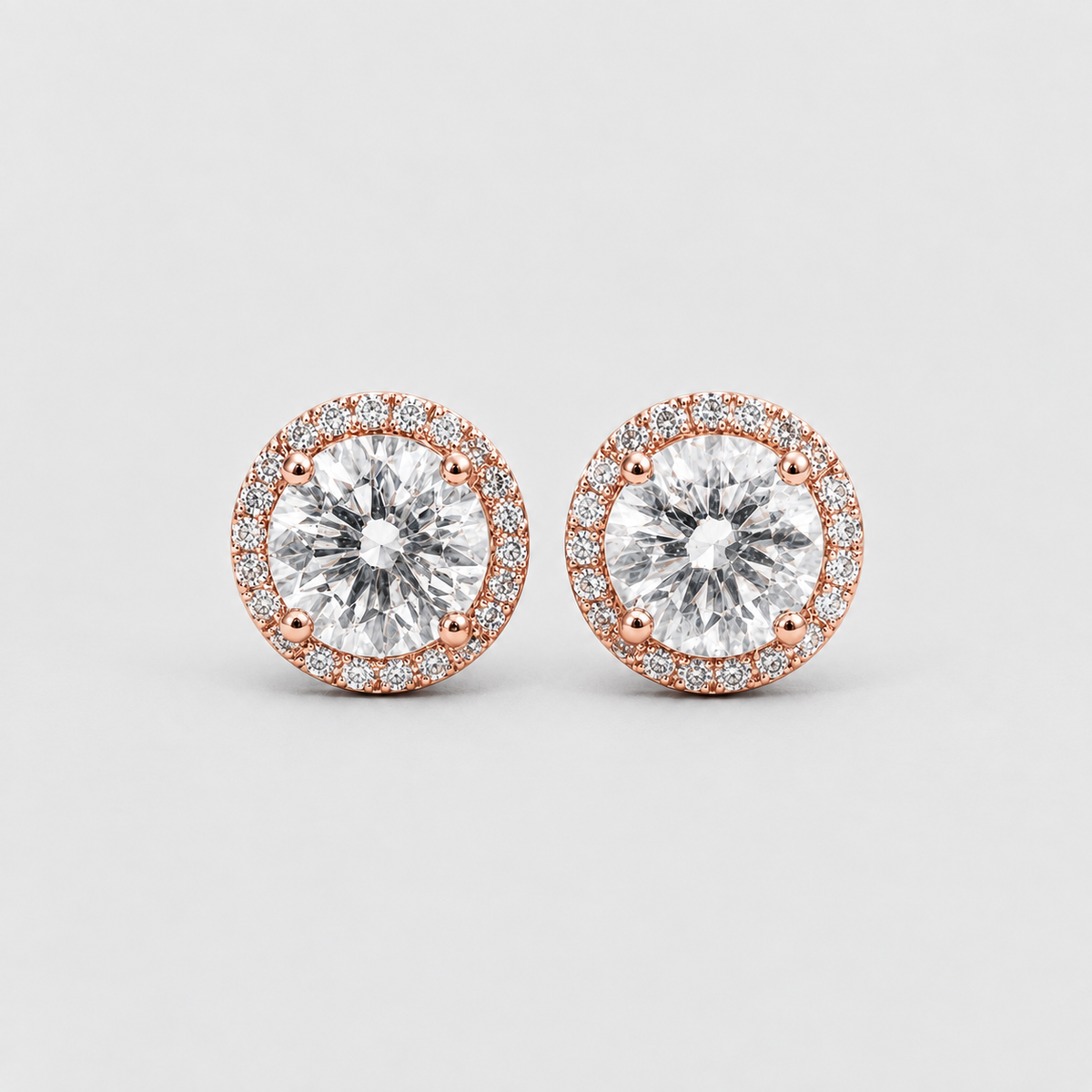 A pair of round halo stud earrings featuring large clear center stones surrounded by a halo of small clear stones in rose gold settings, shown on a light gray background.