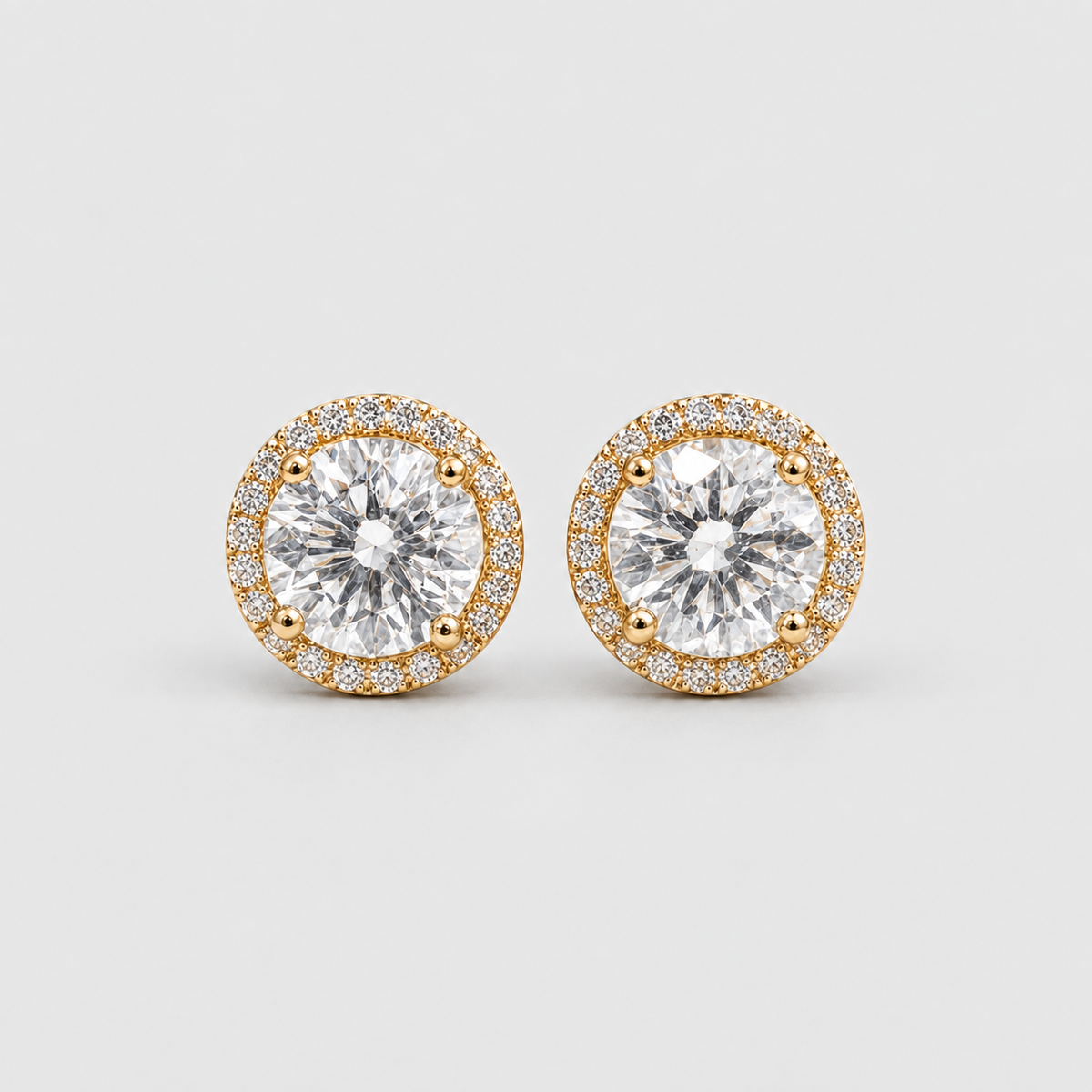 A pair of round stud earrings featuring large clear center stones surrounded by a halo of small clear stones in a yellow gold setting, photographed on a light gray background.