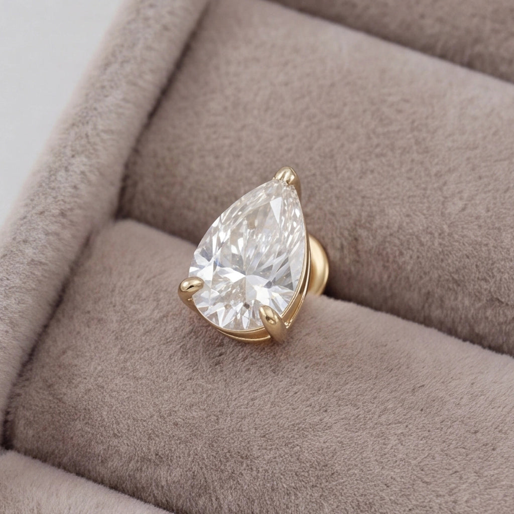 A pear-cut clear gemstone ring in a gold prong setting, displayed on a soft taupe velvet jewelry box.