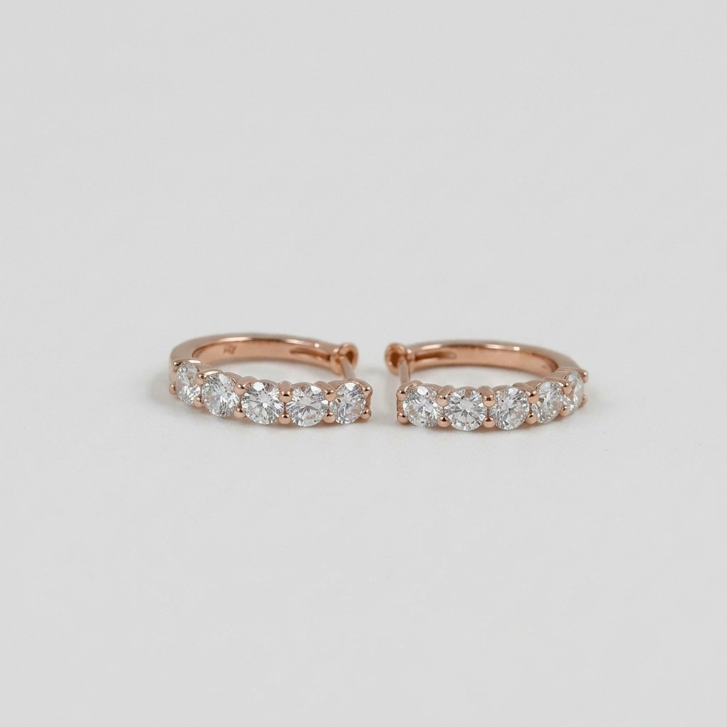 A pair of rose gold huggie hoop earrings with a row of round-cut clear gemstones, photographed on a clean light gray background.
