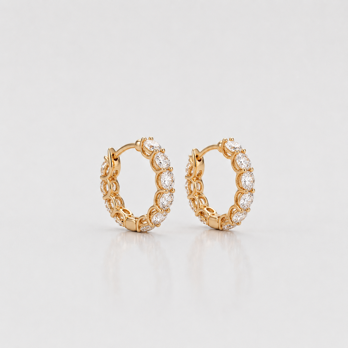 A pair of gold huggie hoop earrings adorned with clear pavé-set round stones, displayed on a clean white reflective surface.