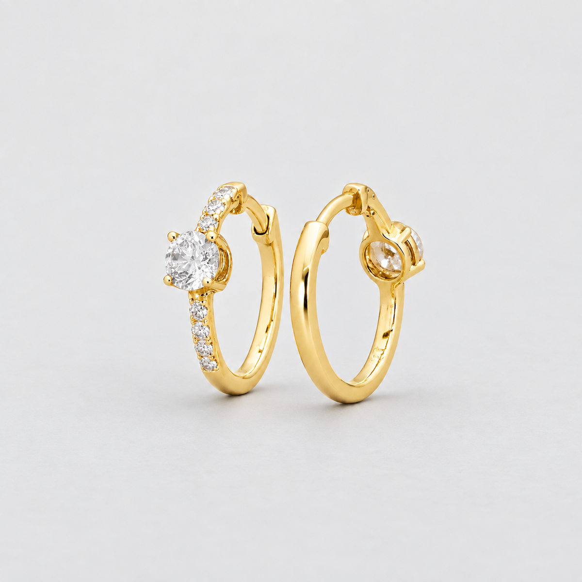 A pair of gold huggie hoop earrings with a round-cut center stone and small pavé-set accent stones, displayed on a light gray background.