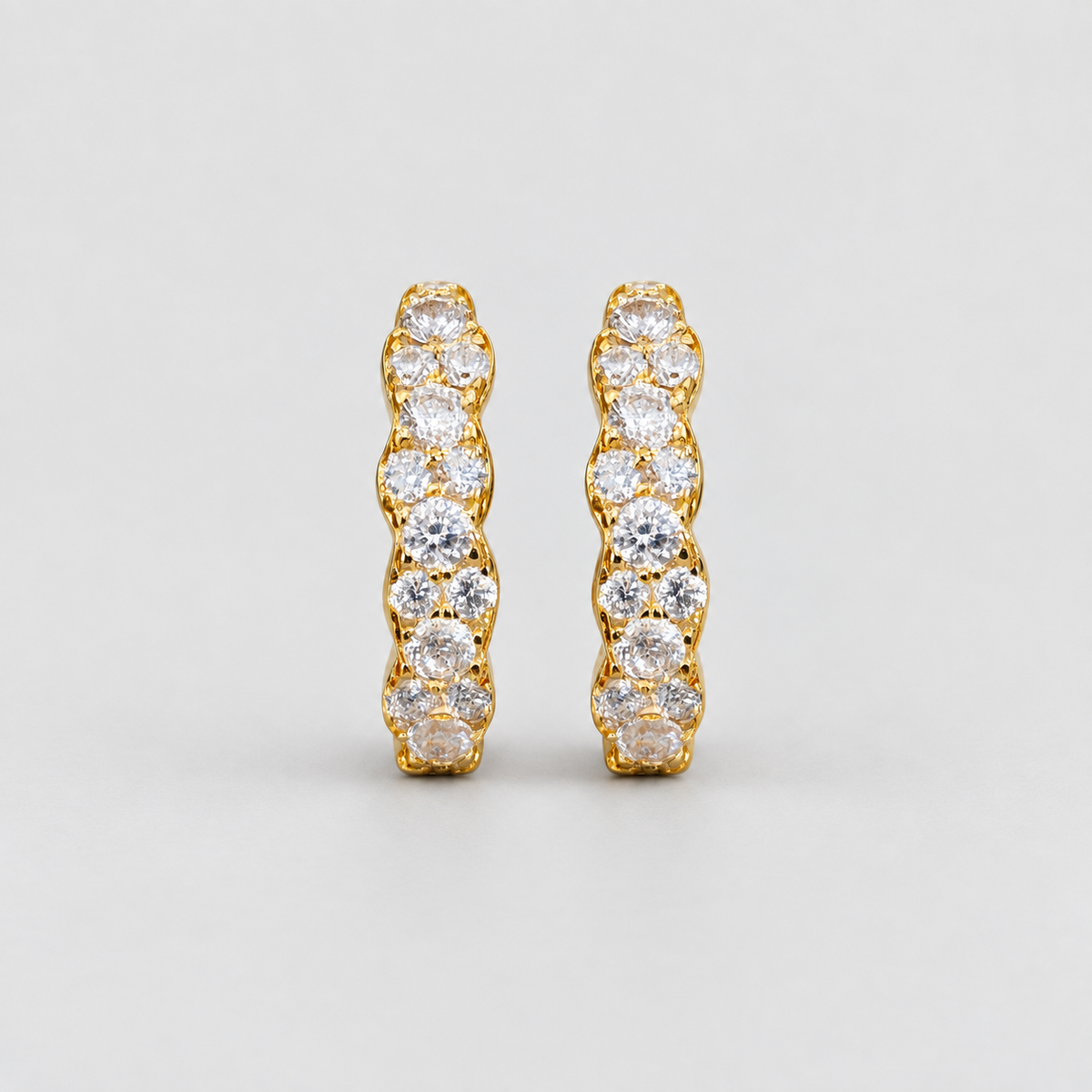A pair of gold huggie hoop earrings set with round-cut clear gemstones, shown against a neutral light gray background.