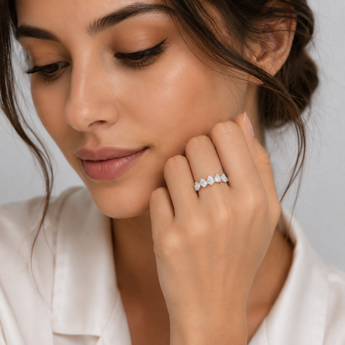 A close-up lifestyle shot of a woman wearing a delicate silver band ring with five small, sparkling heart-shaped clear gemstones. She is wearing a white satin shirt, and the background is a soft, neutral gray.