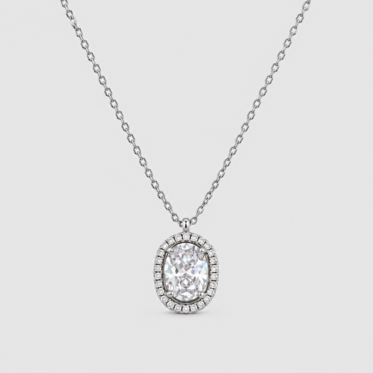 An oval halo pendant necklace featuring a brilliant-cut center stone surrounded by a halo of small pavé-set stones, suspended from a delicate silver-toned chain against a light gray background.
