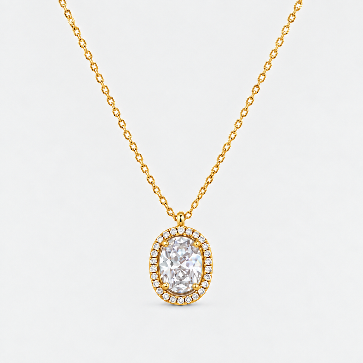 A gold chain necklace with an oval pendant featuring a large clear central stone surrounded by a halo of small clear stones, shown on a light gray background.
