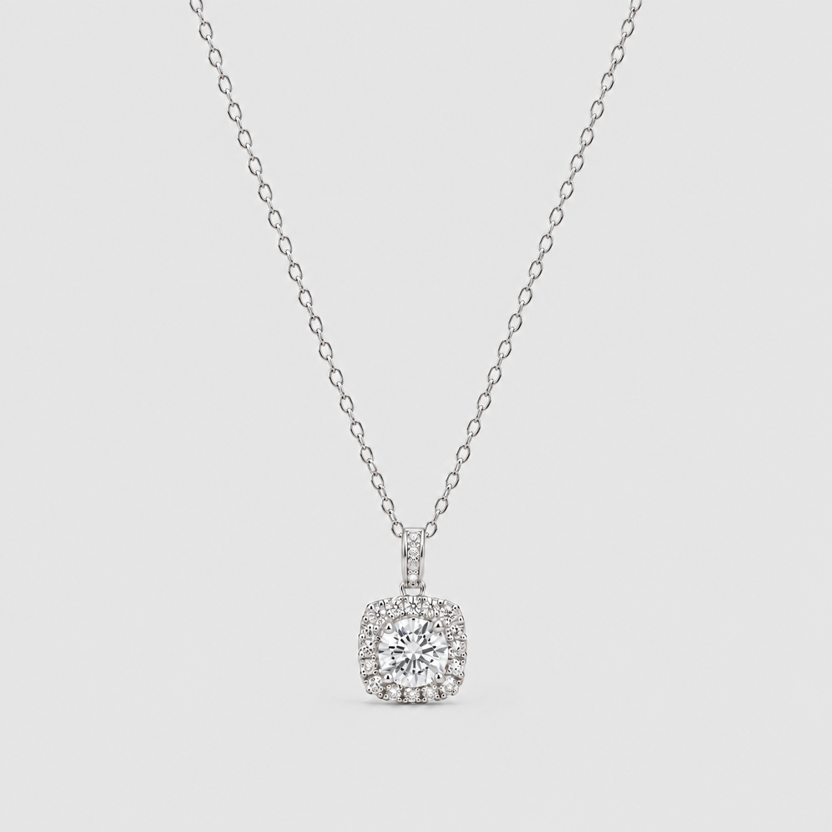 A silver-toned diamond necklace featuring a cushion-cut halo pendant with a central round diamond surrounded by a halo of smaller diamonds, suspended from a delicate cable chain against a light gray background.