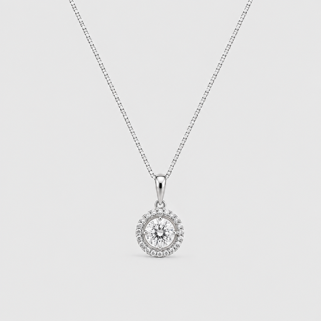 A silver-toned necklace with a box chain and a round pendant featuring a central clear gemstone surrounded by a halo of smaller stones, shown on a light gray background.
