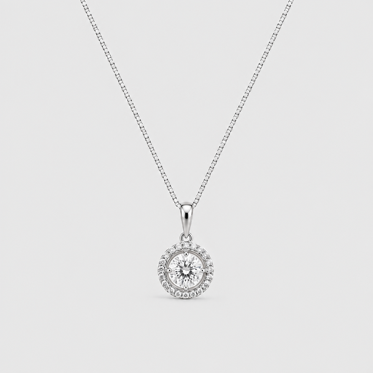 A silver-toned necklace with a box chain and a round pendant featuring a central clear gemstone surrounded by a halo of smaller stones, shown on a light gray background.