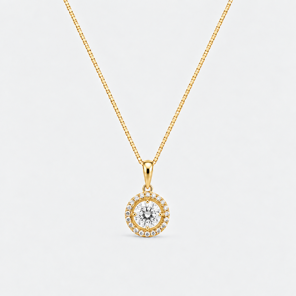 A gold pendant necklace featuring a central round clear gemstone surrounded by a halo of small clear stones, suspended from a gold box chain against a light gray background.