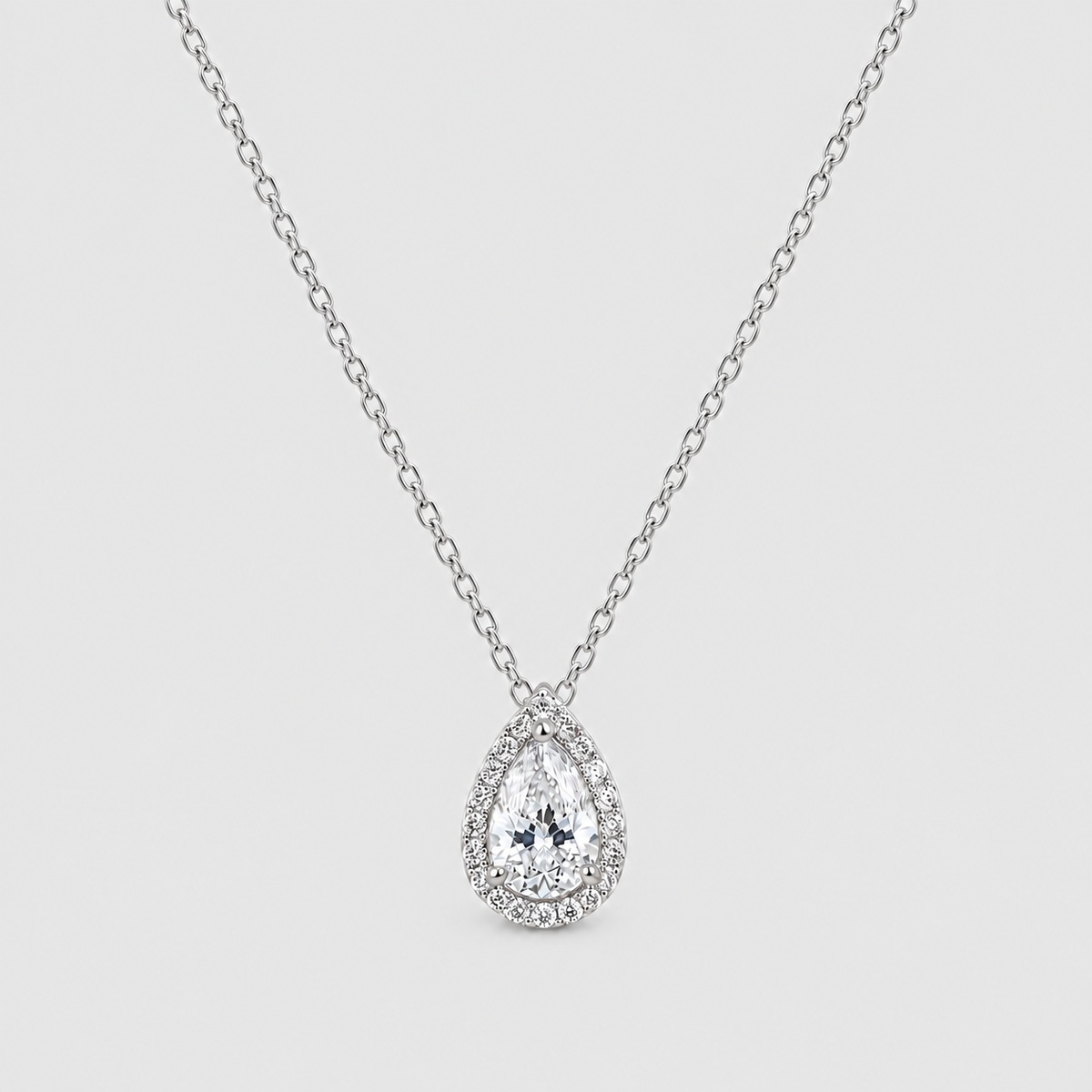 A silver-tone pendant necklace featuring a teardrop-shaped clear gemstone surrounded by a halo of smaller round stones, suspended from a delicate link chain against a light gray background.