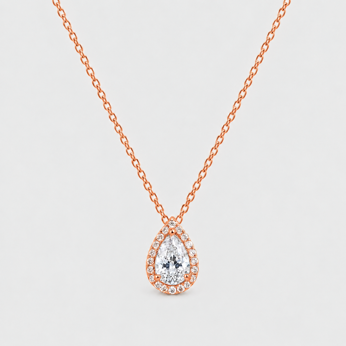 A pear-shaped diamond pendant necklace in rose gold, featuring a halo of small diamonds around the main teardrop-cut center stone, suspended from a delicate rose gold chain against a light gray background.