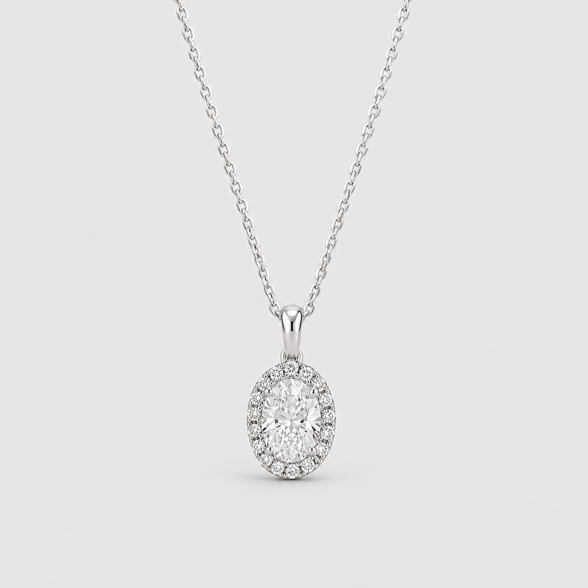 An oval halo pendant necklace featuring a central round-cut diamond surrounded by a halo of smaller diamonds, suspended from a delicate silver-toned chain against a light gray background.