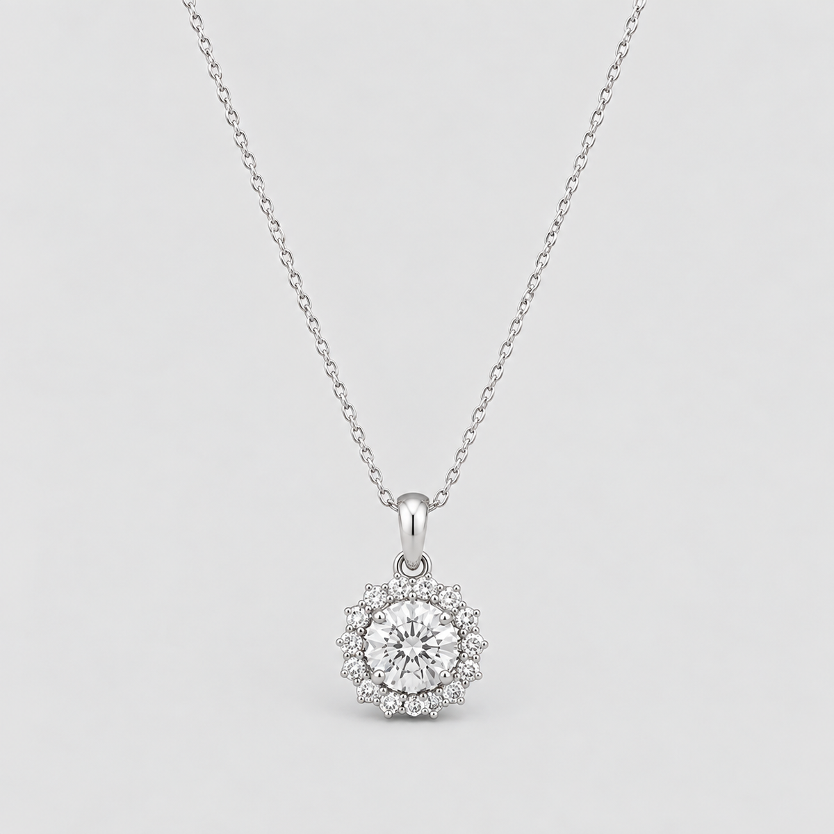 A silver pendant necklace featuring a round-cut clear center stone surrounded by a halo of smaller stones, suspended from a delicate silver chain against a light gray background.