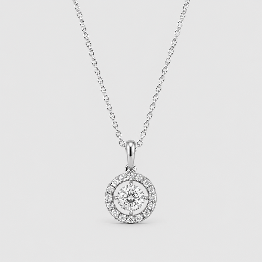 A silver pendant necklace featuring a round halo design with a central clear gemstone surrounded by a halo of smaller stones, suspended from a delicate chain against a light gray background.