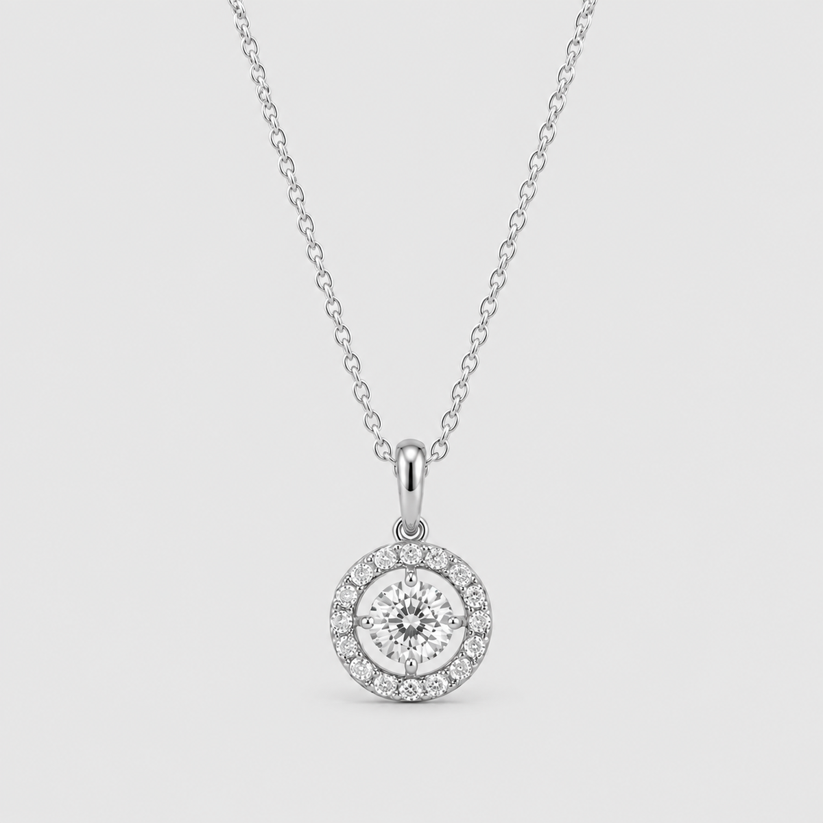A silver pendant necklace featuring a round halo design with a central clear gemstone surrounded by a halo of smaller stones, suspended from a delicate chain against a light gray background.