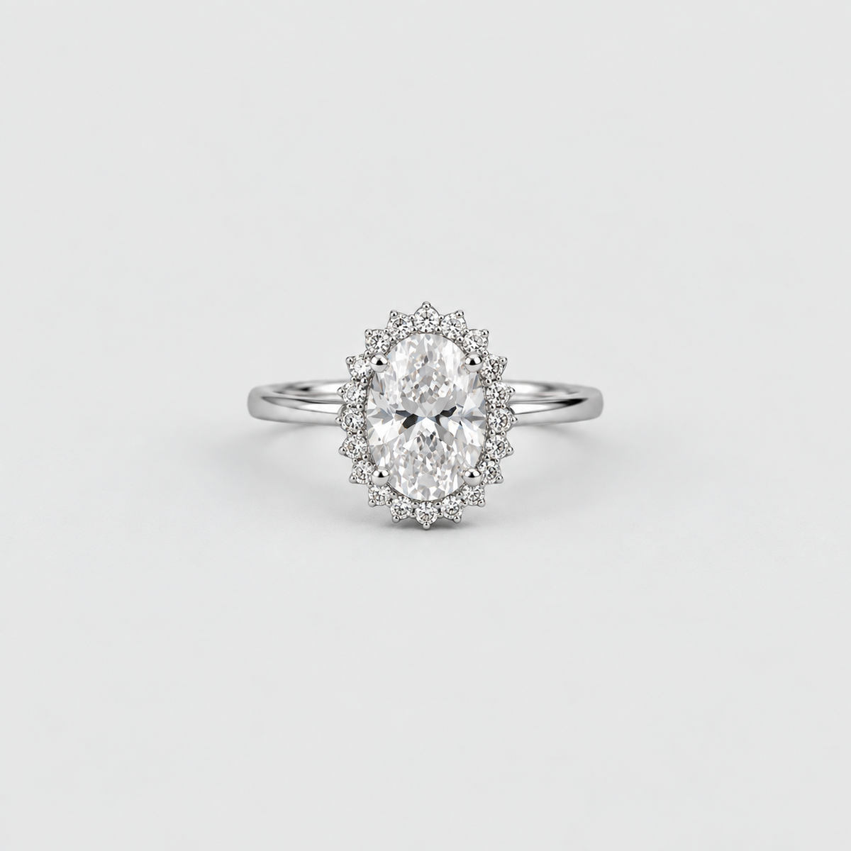 An oval halo ring featuring a brilliant-cut center stone surrounded by a double halo of small round stones, set on a polished silver-colored band, shot against a seamless light gray background.