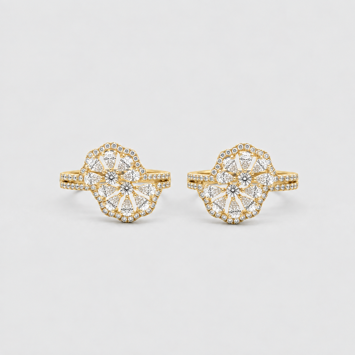 A pair of gold rings with large, intricate floral cluster settings paved with clear stones, photographed on a clean, light gray background.