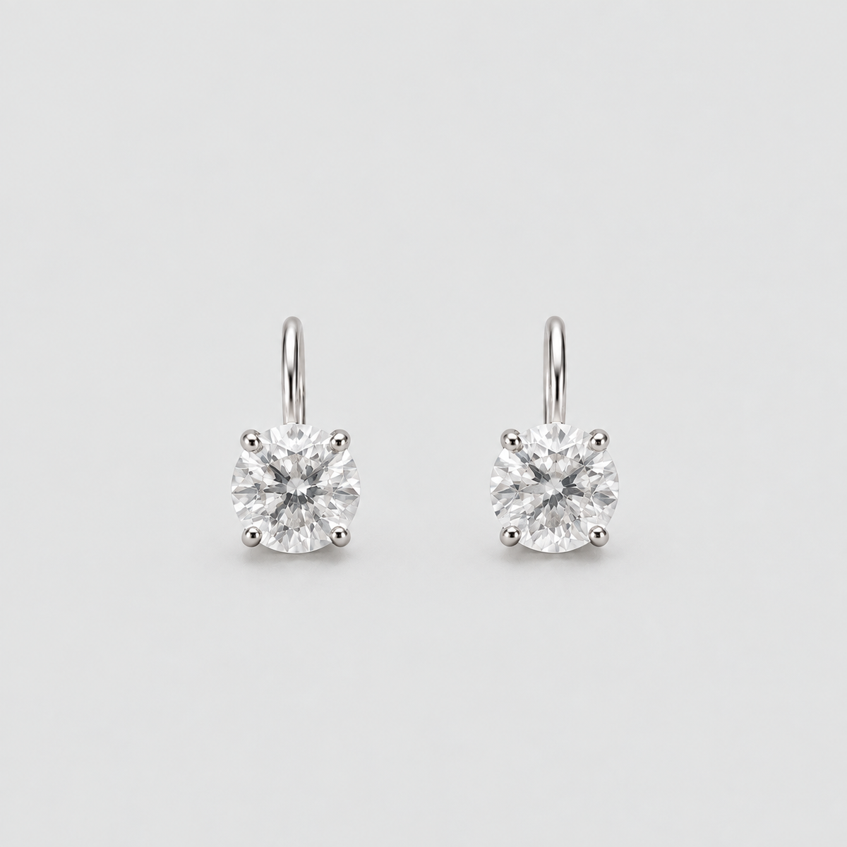 A pair of solitaire drop earrings with round brilliant-cut clear gemstones in a silver-toned four-prong setting, suspended from simple silver-toned hooks against a light gray background.