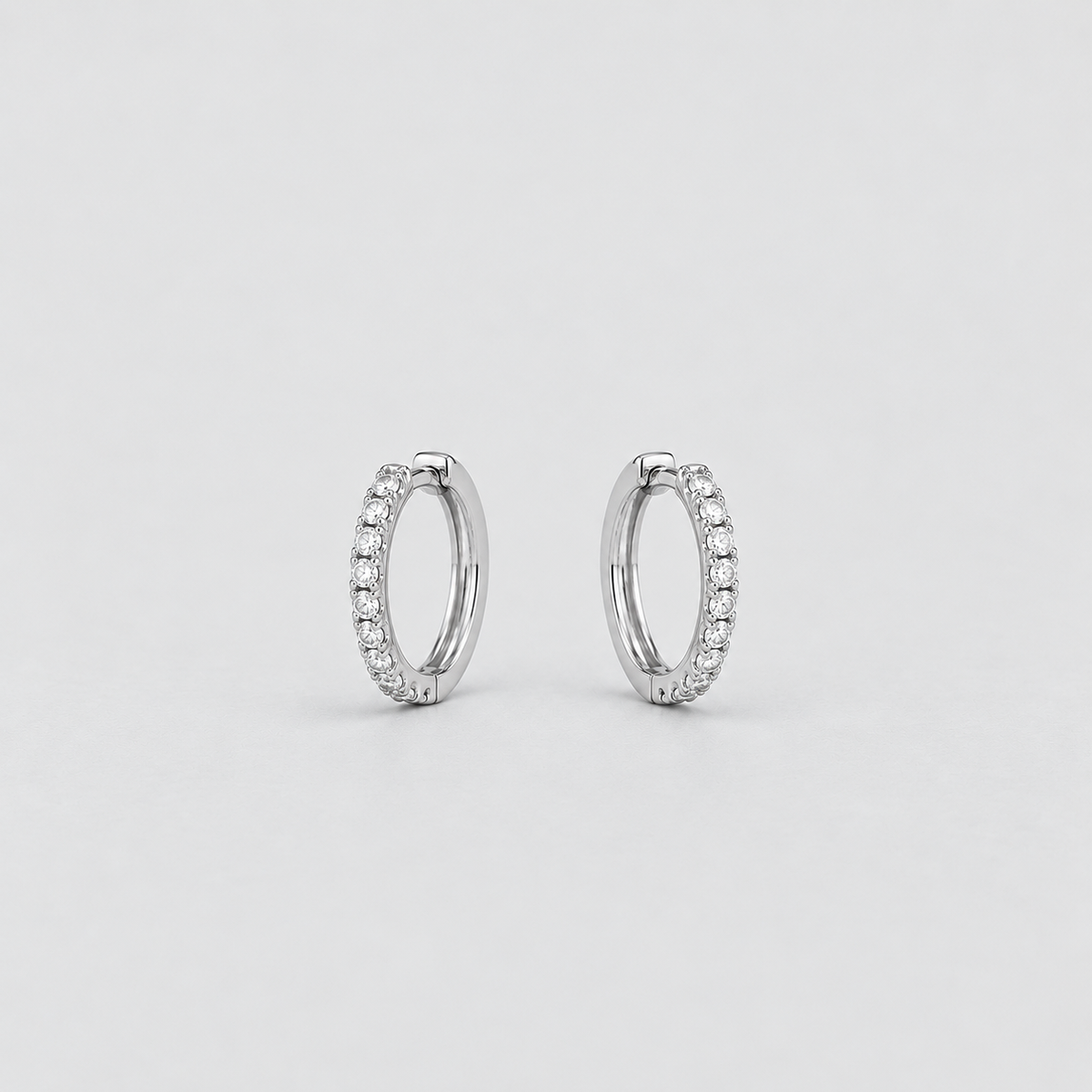 A pair of silver-toned huggie hoop earrings adorned with small clear pavé stones, set against a clean, light gray background.