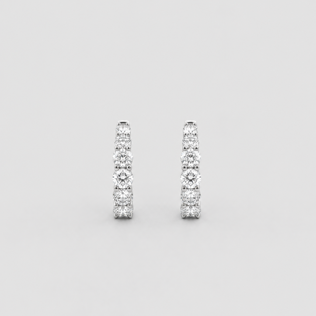 A pair of silver-toned huggie hoop earrings set with round-cut clear gemstones, shown against a solid light gray background.