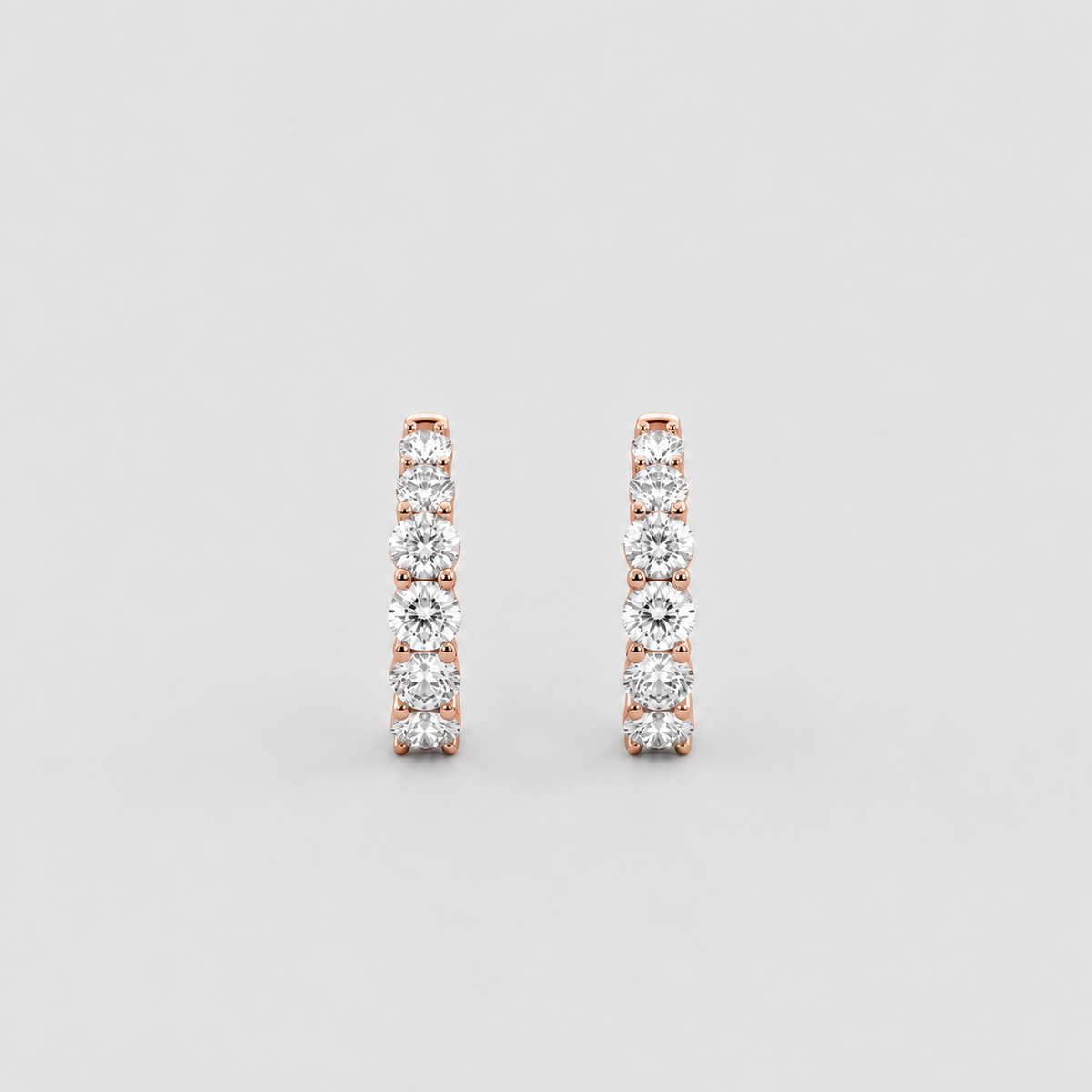 A pair of rose gold huggie hoop earrings set with round-cut clear gemstones, displayed against a neutral light gray background.
