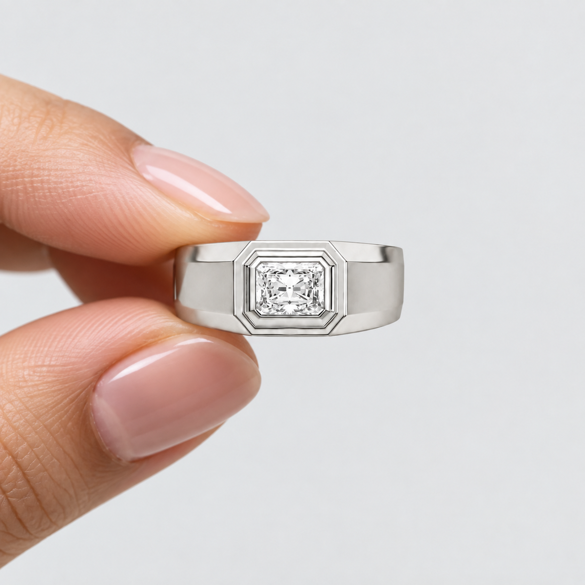 A silver-toned men's ring with a square bezel-set diamond, held between fingers against a light gray background.