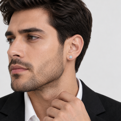 A close-up of a man wearing a small round brilliant-cut diamond stud earring on his earlobe, dressed in a black suit jacket and white collared shirt against a light gray background.