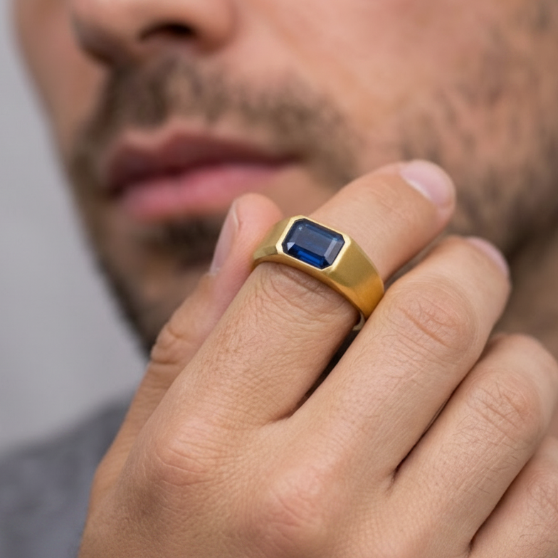 A close-up of a man's hand wearing a gold signet ring with a rectangular blue gemstone, with his face blurred in the background.