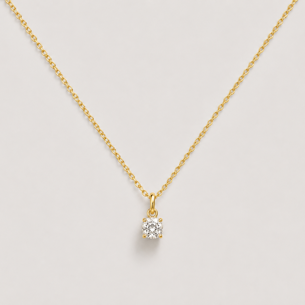 A delicate gold chain necklace with a small, clear gemstone pendant set in a gold bezel, displayed against a light gray background.