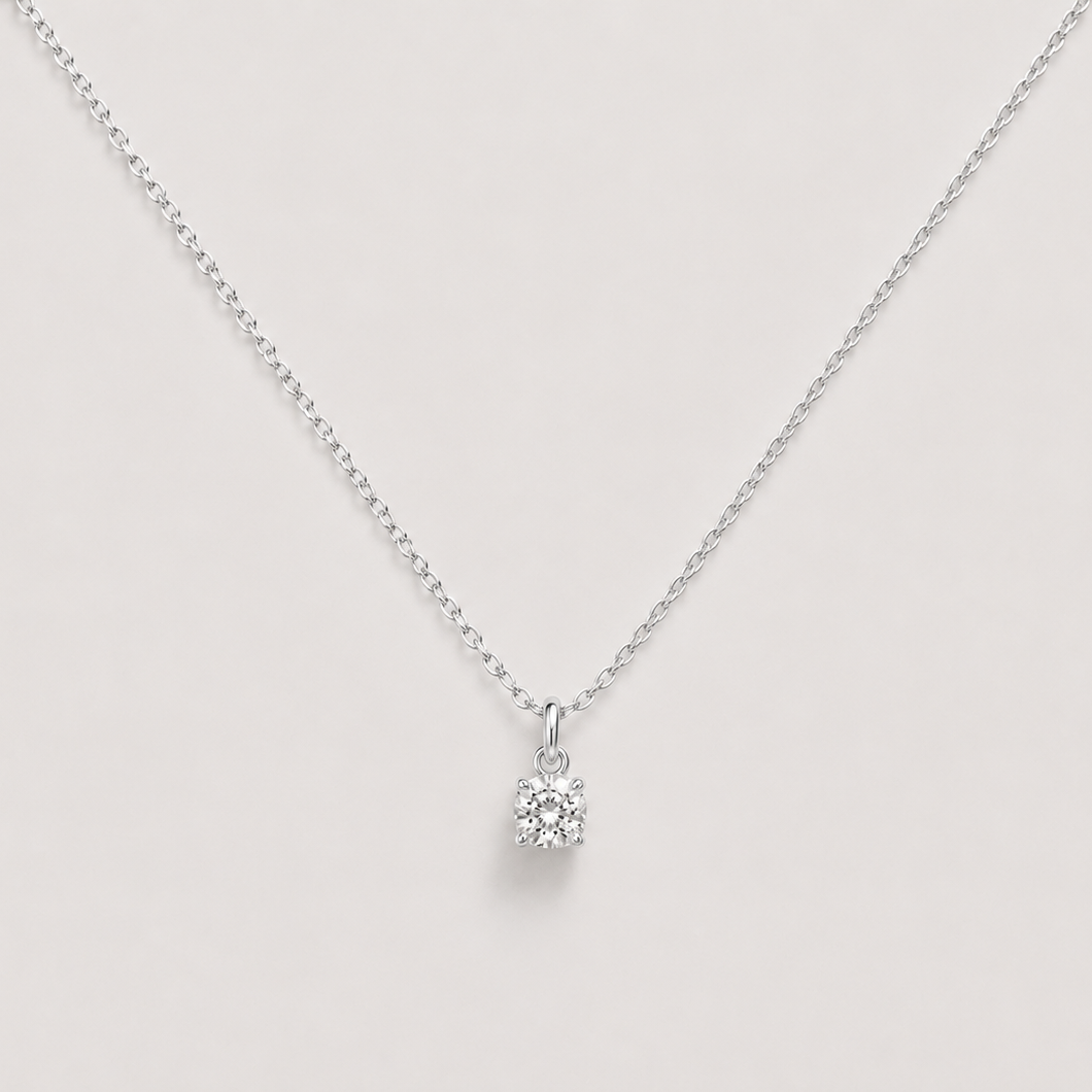 A delicate silver chain necklace with a small, clear gemstone pendant set in a silver bezel, displayed against a solid light beige background.