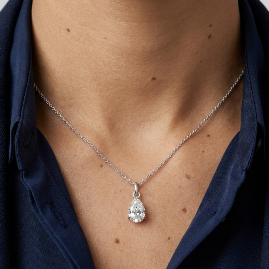 A close-up of a person wearing a delicate silver chain necklace with a teardrop-shaped clear gemstone pendant, styled with a dark navy blue collared shirt.