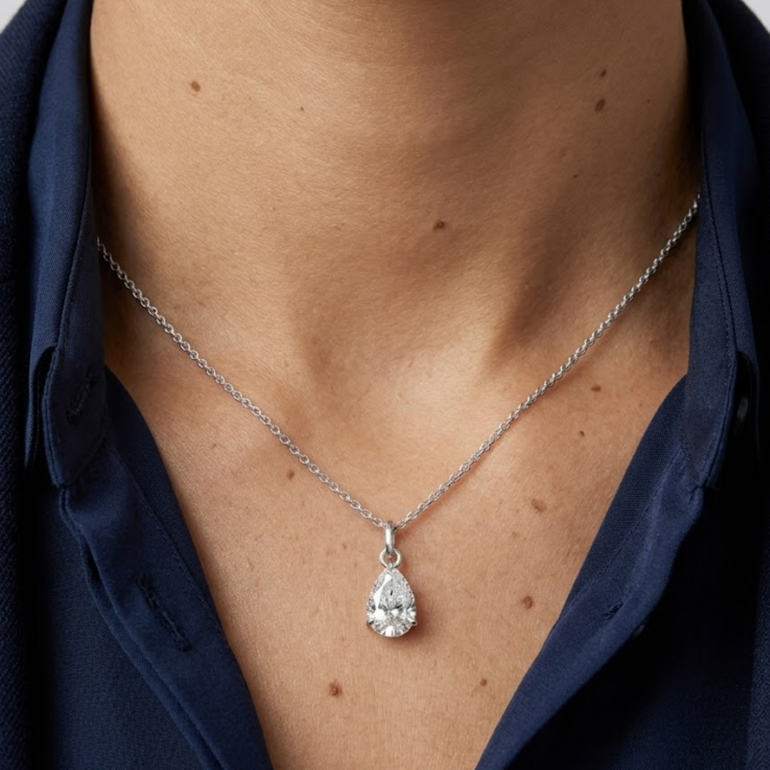 A close-up of a person wearing a delicate silver chain necklace with a teardrop-shaped clear gemstone pendant, styled with a dark navy blue collared shirt.