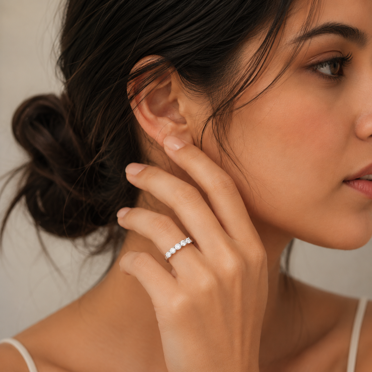 A close-up lifestyle shot of a woman wearing a delicate silver band ring with small round clear stones on her finger, with her hand resting near her ear.