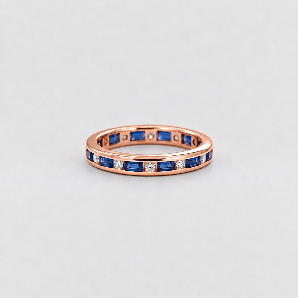 A rose gold eternity ring featuring baguette-cut blue sapphires and small round-cut clear stones, set against a clean light gray background.
