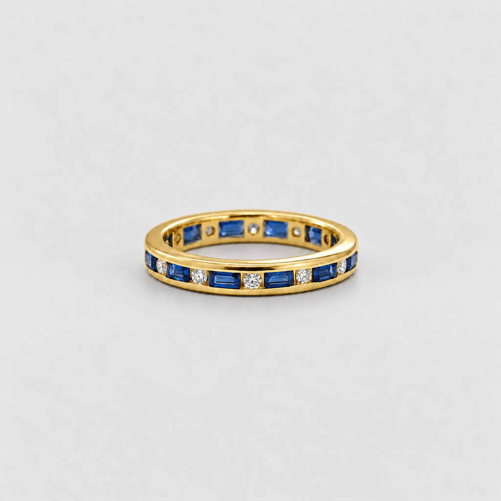 A yellow gold eternity ring featuring baguette-cut blue sapphires and small round-cut clear stones, shown on a neutral light gray background.