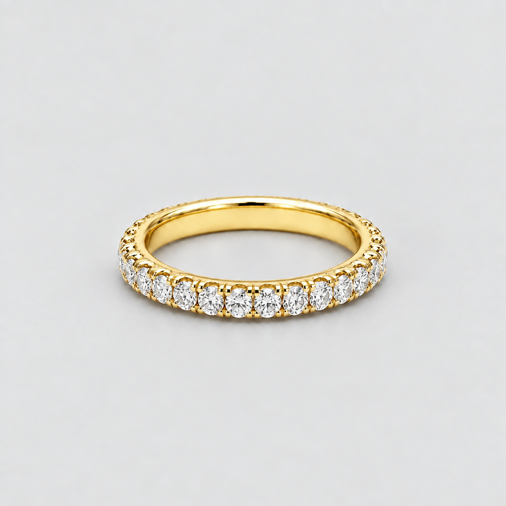 A yellow gold eternity ring featuring a continuous row of round-cut clear gemstones in a prong setting, shown on a neutral light gray background.