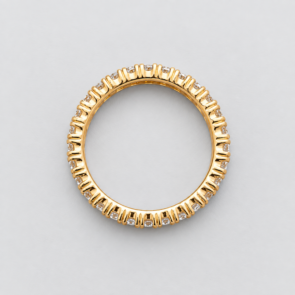 A yellow gold eternity ring featuring a continuous line of small, round-cut clear gemstones in a prong setting, photographed from directly above on a neutral light gray background.