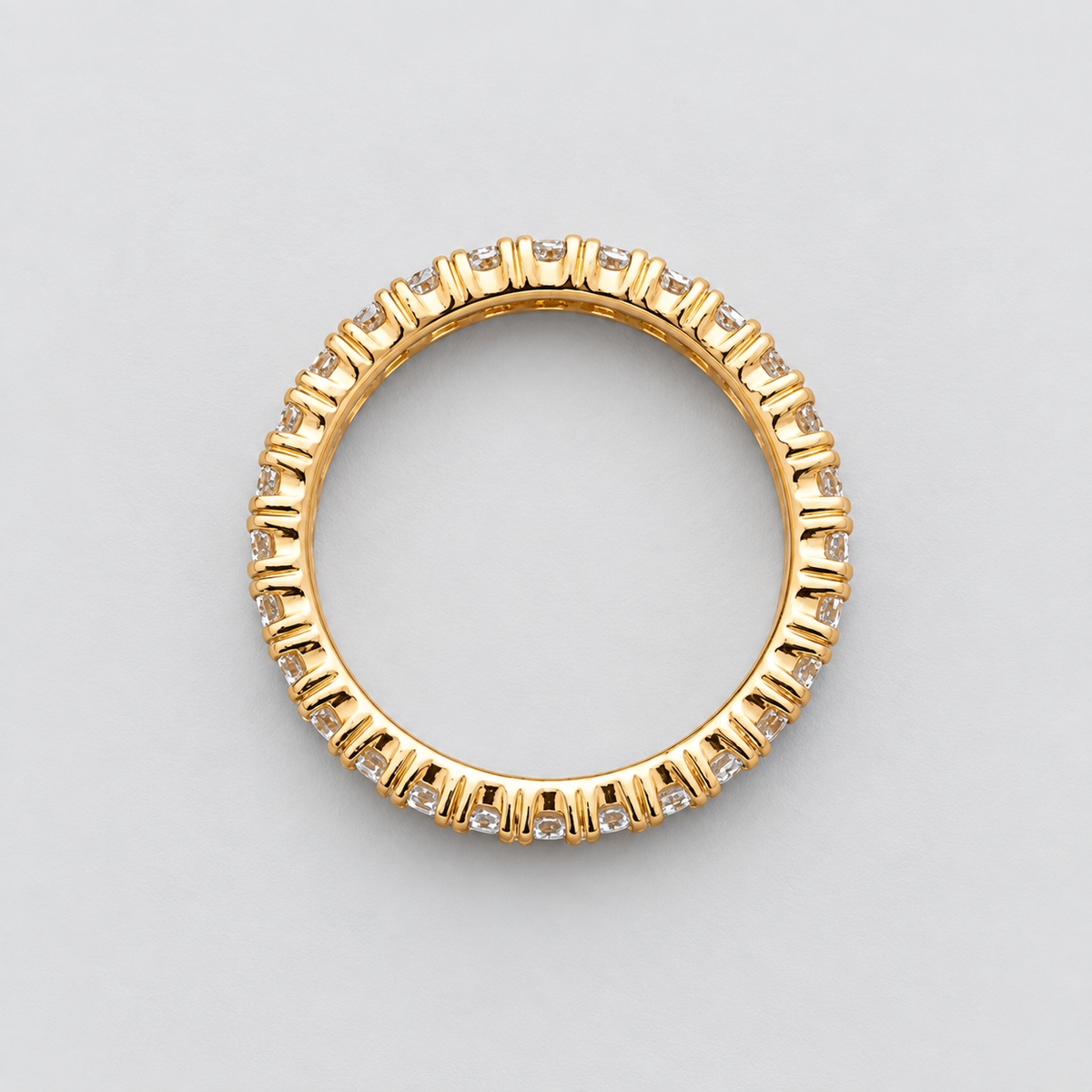 A yellow gold eternity ring featuring a continuous line of small, round-cut clear gemstones in a prong setting, photographed from directly above on a neutral light gray background.