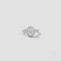 A halo ring featuring a round-cut center stone surrounded by a halo of smaller stones, set on a silver-toned pavé band, photographed on a light gray background.
