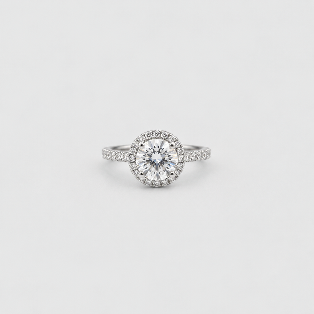 A silver-toned halo ring featuring a round brilliant-cut center stone surrounded by a halo of smaller stones, with a pavé-set band, shown on a light gray background.