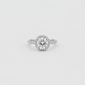 A silver-toned halo ring featuring a round brilliant-cut center stone surrounded by a halo of smaller stones, with a pavé-set band, shown on a light gray background.