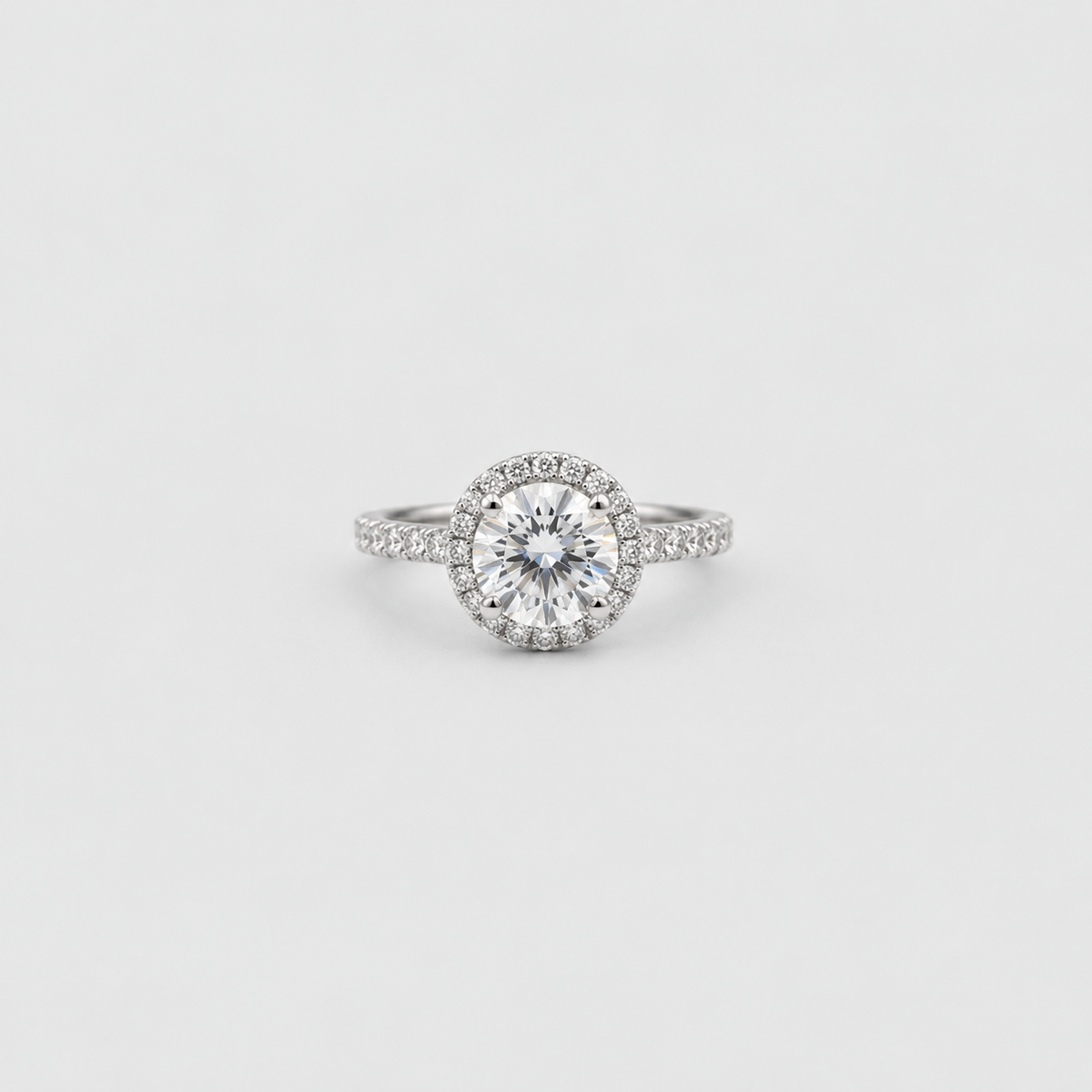 A silver-toned halo ring featuring a round brilliant-cut center stone surrounded by a halo of smaller stones, with a pavé-set band, shown on a light gray background.