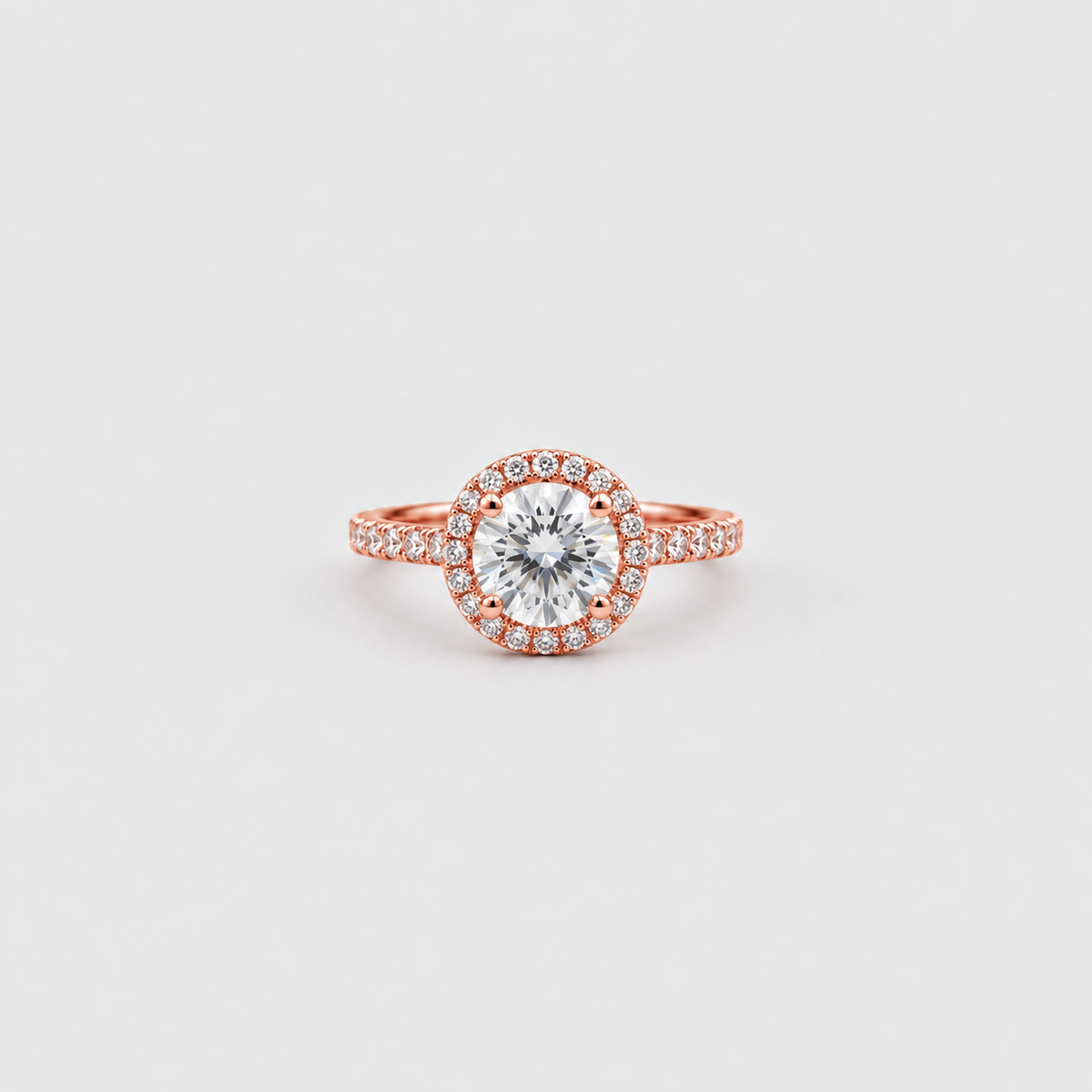 A rose gold halo ring featuring a round-cut clear center stone surrounded by a halo of small pavé-set stones, with a matching halo around a smaller center stone, set against a light gray background.