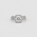 A silver-toned halo ring featuring a round brilliant-cut center stone surrounded by a halo of smaller stones, with a three-stone split band also paved with small diamonds, set against a light gray background.