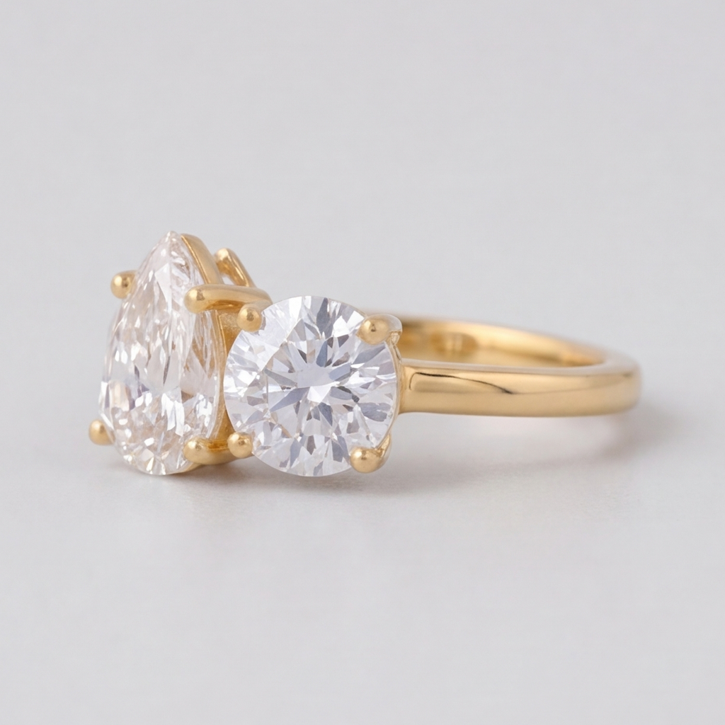 A yellow gold ring featuring a pear-cut diamond on the left and a round brilliant-cut diamond on the right, set on a polished gold band against a light gray background.